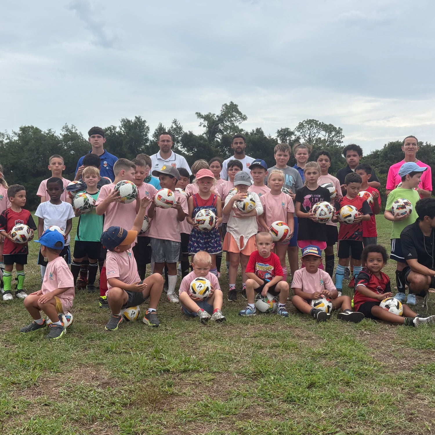 Group of children and adults holding soccer balls on a grassy field with trees in the background.