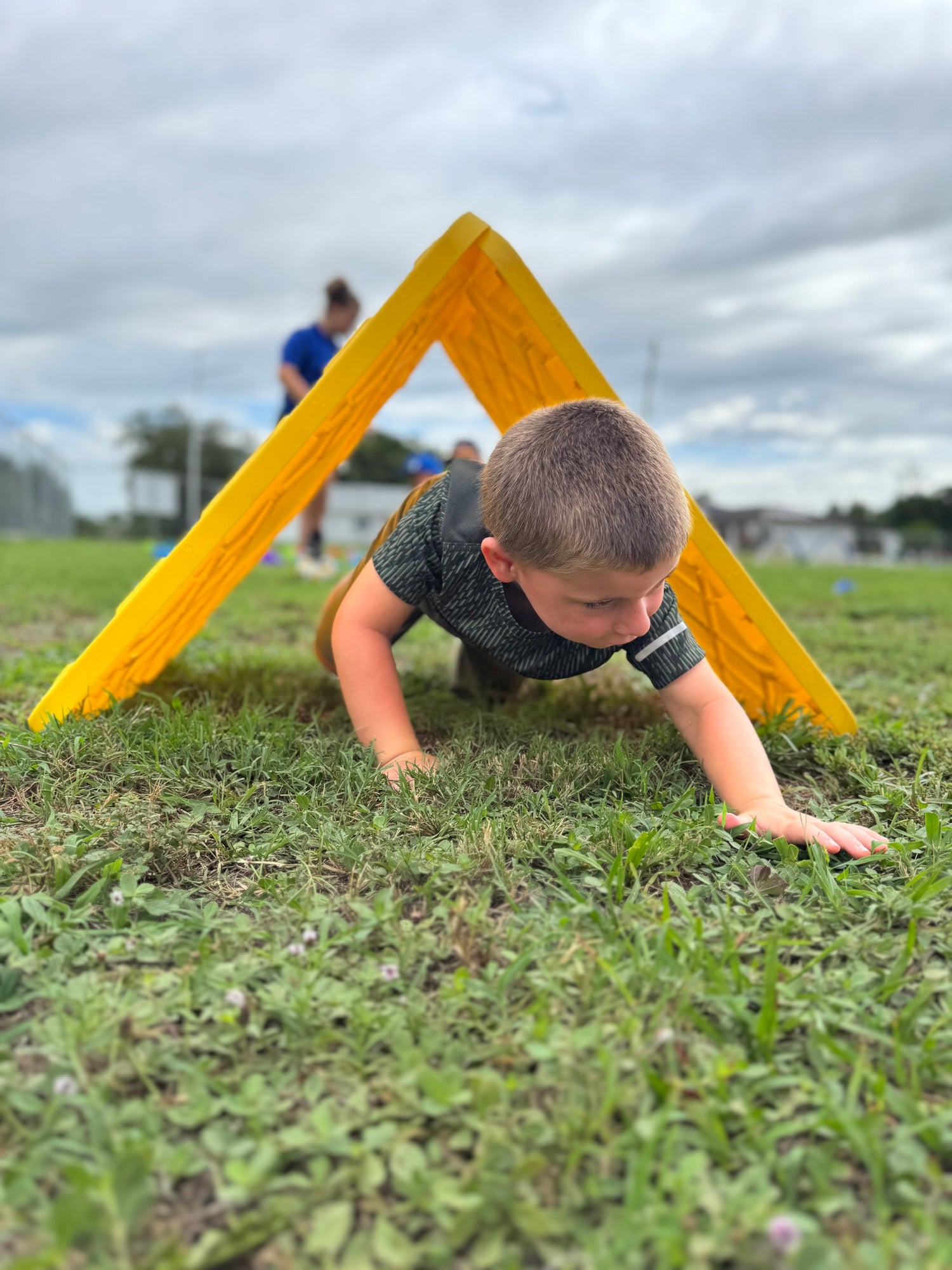 A young boy crawling under a yellow A-frame wooden obstacle on a grassy field, with a cloudy sky in the background.