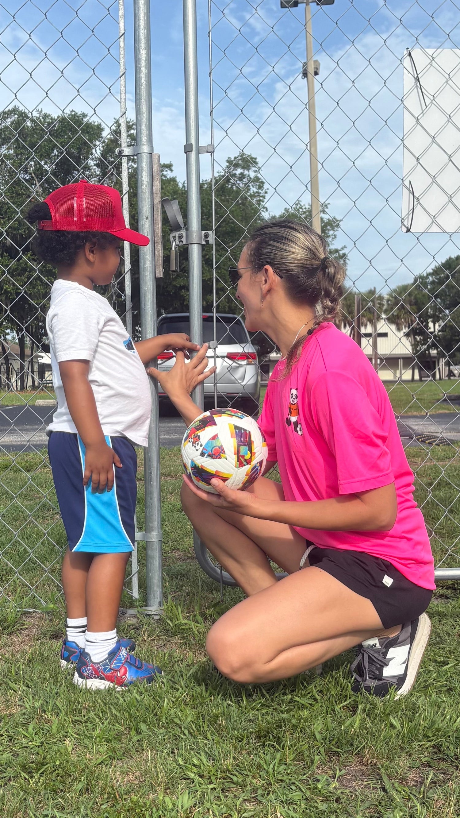 Woman in pink shirt and black shorts kneeling on grass, interacting with a child wearing a red cap and white shirt, with a soccer ball between them, in an outdoor setting.